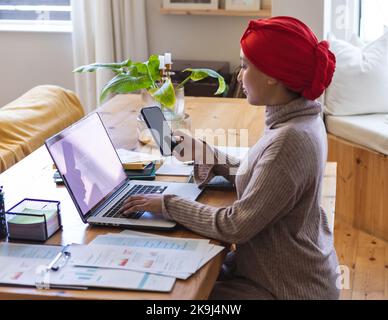 Biracial woman wearing hijab, sitting at table in living room and using laptop with copy space Stock Photo