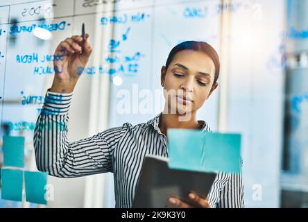 Step one to success is proper planning. a young businesswoman using a digital tablet while writing notes on a glass wall in an office. Stock Photo