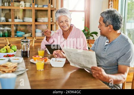 Enjoying your breakfast. a carefree elderly couple having breakfast together while reading the paper and browsing on a digital tablet at home. Stock Photo