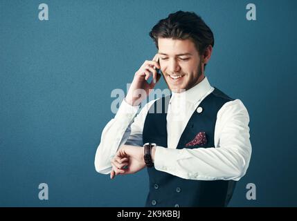 I can be there in 25 minutes. Studio shot of a handsome young businessman checking the time while making a phonecall against a blue background. Stock Photo