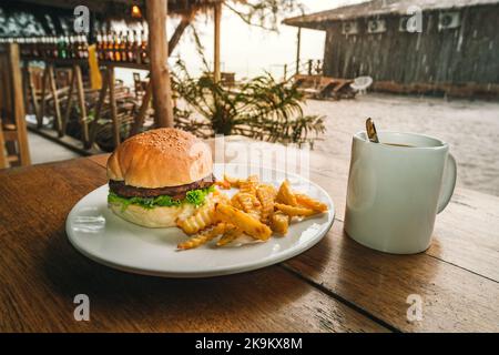 Big juicy Burger and fries on a white plate with a Cup of coffee. American Breakfast at an outdoor cafe on the background of a tropical landscape. fas Stock Photo