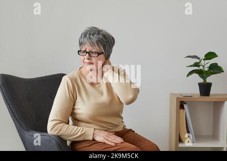 Unhappy older woman feels pain in her neck and shoulders while sitting on chair at home. Stock Photo