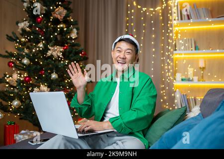 Christmas man at home on sofa smiling and looking at camera, Asian man near Christmas tree with laptop holding hand up greeting gesture using laptop for remote work and new year video call. Stock Photo