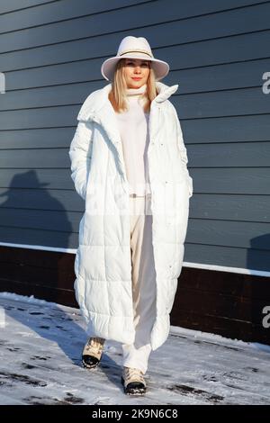 Young woman posing in total white fashion sport chic look with long down jacket, wool costume and fedora. Fashionable blonde model, winter street styl Stock Photo
