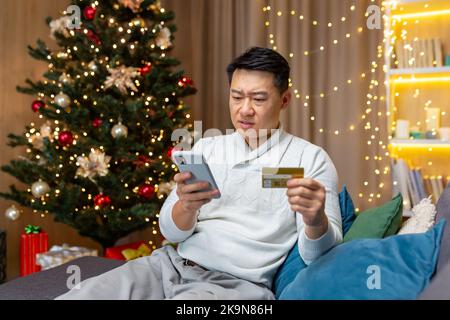 Dissatisfied Asian man trying to make a purchase in an online internet store, man cheated on Christmas, sitting on sofa in living room with smartphone in hands and credit card at home near Christmas tree Stock Photo