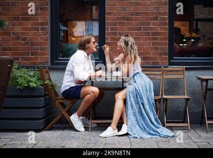 fashionable beautiful Young couple sitting in a cafe and drinking coffee outside near the house. natural color. stylish woman feeding with spoon man i Stock Photo