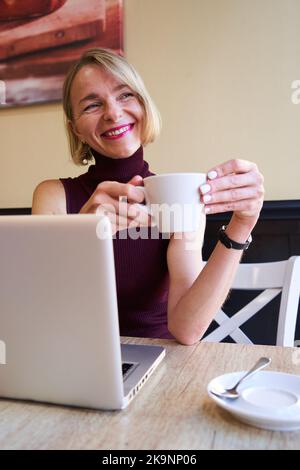Vertical photo of a mid adult woman working from a cafeteria and drinking coffee while daydreaming. Stock Photo