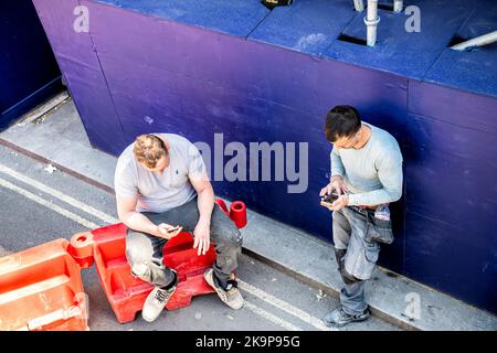 London, United Kingdom - June 22, 2018: Two construction workers men taking lunch break, using phones outside on street of city of Westminster Stock Photo