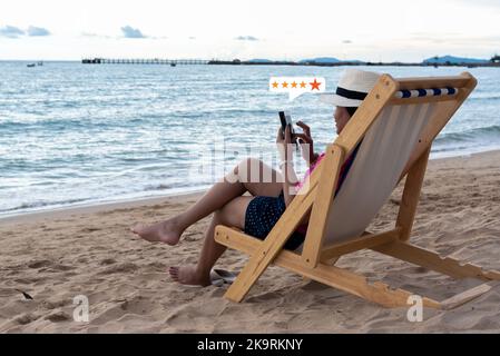 Young Asian happy woman using smartphone on the beach ,customer service and five stars satisfaction rating. Stock Photo