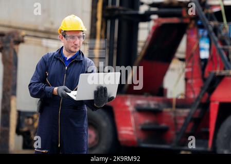 Young worker engineer wearing yellow safety hard hat using laptop computer laptop to checking and control loading containers box from cargo ship for i Stock Photo