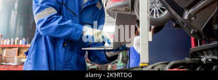 Young man is mechanic using laptop computer checking engine of car in the garage, worker examining and diagnostic automobile, technician and maintenan Stock Photo