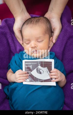 newborn baby holding ultrasound copy in hand and sleeping at mother palm in baby wrap Stock Photo