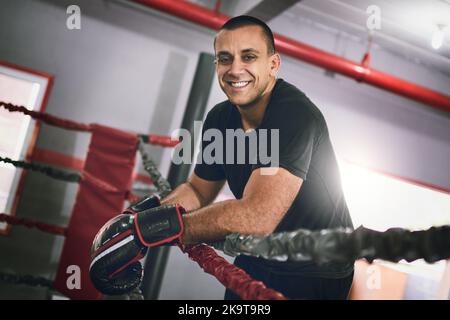 You may take a break now. Portrait of a cheerful young male boxer leaning against ropes inside of a boxing ring at a gym during the day. Stock Photo