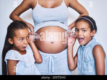 I heard something. Portrait of two cheerful little girls standing next their mother while each putting a glass on her pregnant belly to listen for