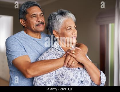 To think we were high school sweethearts. a cheerful mature couple holding each other while looking outside through a window at home during the day. Stock Photo