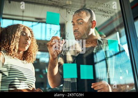 Their minds think together as one. two focussed young work colleagues putting up sticky notes on a glass wall while brainstorming together in the Stock Photo