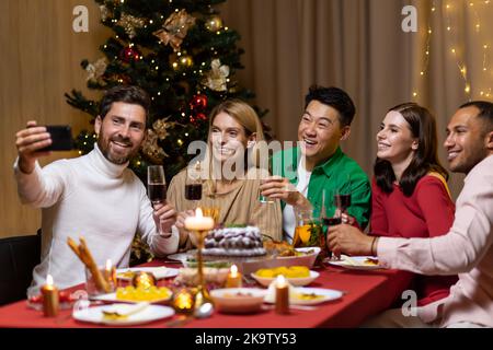 Diverse friends celebrating Christmas and New Year in the evening, sitting at the table, drinking wine and eating, taking a selfie together on the phone near the Christmas tree. Stock Photo