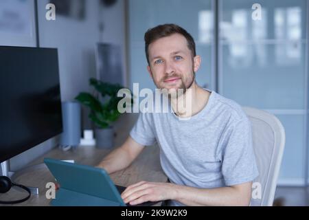 Smart tech concept. Attractive bearded male sitting in home office using tablet. Close-up of portrait of young handsome man in grey shirt and looking in camera, positive emotion. High quality photo Stock Photo