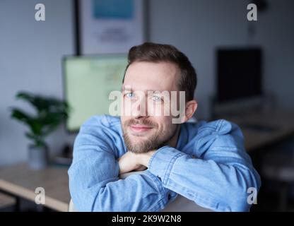 Attractive bearded male sitting in home office looking at camera smiling. Young handsome man in denim shirt, positive emotion with computer monitor on background. High quality image Stock Photo