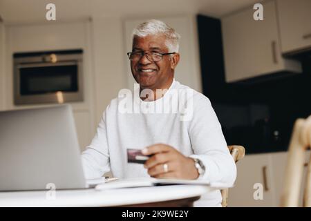 Happy senior man smiling at the camera while shopping online at home. Mature man using his credit card to place an online order on a laptop. Cheerful Stock Photo