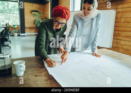 Cheerful Muslim architects working on blueprint drawings in a modern office. Two creative businesswomen planning a new innovative project. Female desi Stock Photo