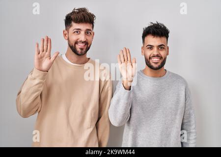 Young homosexual couple standing over white background waiving saying hello happy and smiling, friendly welcome gesture Stock Photo