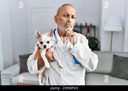 Mature veterinarian man checking dog health serious face thinking about question with hand on chin, thoughtful about confusing idea Stock Photo