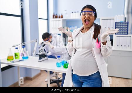Young hispanic woman expecting a baby working at scientist laboratory celebrating victory with happy smile and winner expression with raised hands Stock Photo