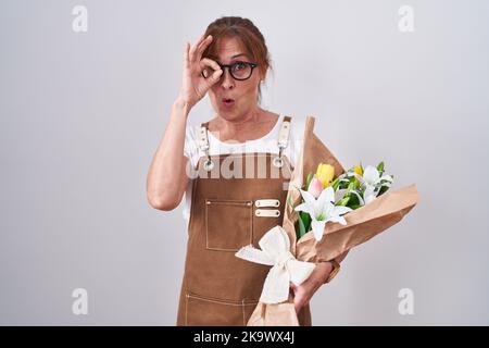 Middle age woman holding bouquet of white flowers doing ok gesture shocked with surprised face, eye looking through fingers. unbelieving expression. Stock Photo