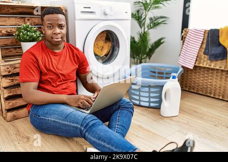 Young african man doing laundry and using computer smiling looking to the side and staring away thinking. Stock Photo