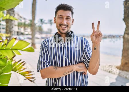 Young handsome man listening to music using headphones outdoors smiling with happy face winking at the camera doing victory sign. number two. Stock Photo