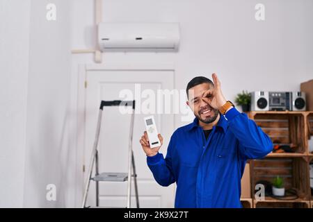 Hispanic repairman working with air conditioner smiling happy doing ok sign with hand on eye looking through fingers Stock Photo