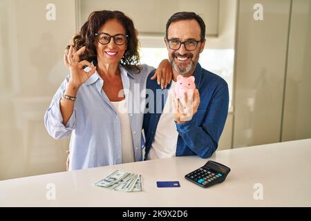 Middle age couple holding piggy bank calculating savings doing ok sign with fingers, smiling friendly gesturing excellent symbol Stock Photo