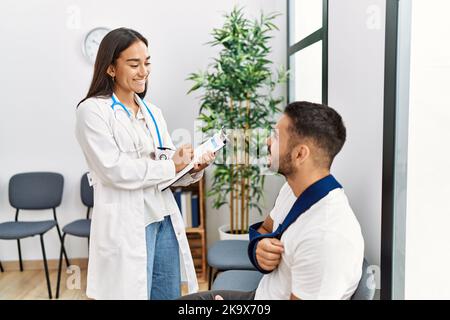 Youg latin injuried man wearing sling talking with the doctor at clinic waiting room. Stock Photo