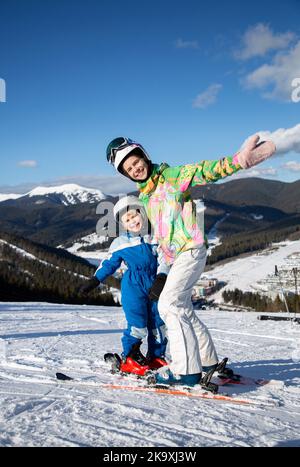 kid boy and teenage girl, brother and sister, are relaxing in ski resort, joyfully standing side by side on skis on top of mountain slope. Winter spor Stock Photo