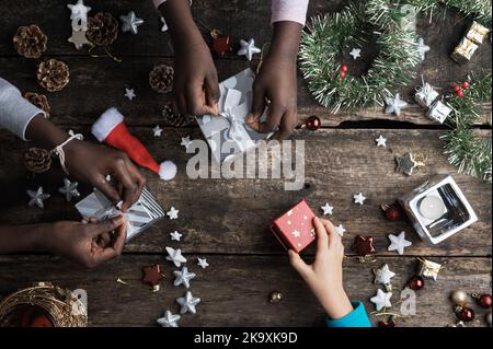 Top view of hand of three children of caucasian and black ethnicities wrapping christmas presents and gift boxes on a rustic wooden desk full of new y Stock Photo