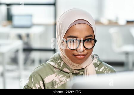Young confident female IT support manager in eyeglasses and hijab sitting in front of computer monitor and networking in office Stock Photo