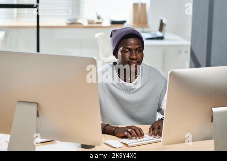 Young black man in casualwear working over new software while sitting in front of computer monitors in openspace office Stock Photo