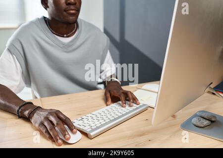 Close-up of young black man in t-shirt clicking on mouse and pressing button of computer keyboard while working in front of monitor Stock Photo