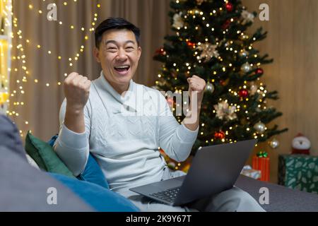 Happy Christmas man looking into camera and smiling celebrating victory and successful triumph at online job, Asian man sitting on sofa at home in living room near Christmas tree. Stock Photo