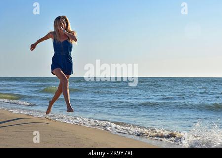 Young blonde woman of white race runs happy along the beach of the seashore in denim overalls Stock Photo
