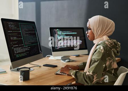 Side view of young Muslim female programmer looking through coded information on computer screen while sitting by workplace Stock Photo
