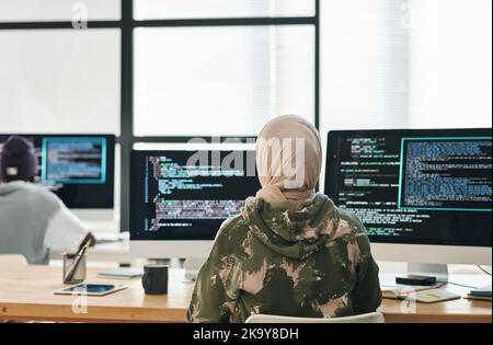 Rear view of young Muslim woman in hijab and casualwear sitting by workplace in front of computer screens with coded data Stock Photo