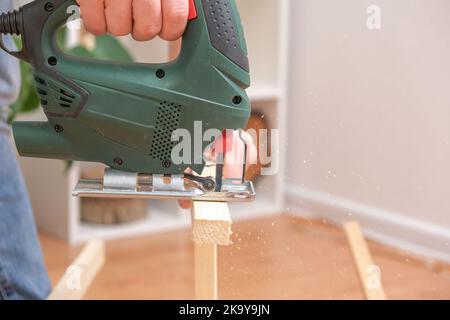 The hands of a master carpenter with an electric jigsaw in his hands cutting off a piece of wood. Male hands using fret saw for cutting wood. A carpenter cuts wood with a jigsaw in a home workshop. Stock Photo