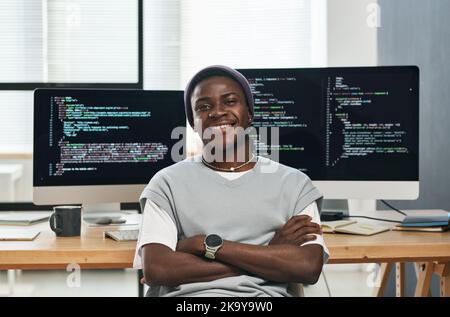 Happy young successful IT engineer in casualwear looking at you while sitting by workplace against computer screens with coded data Stock Photo