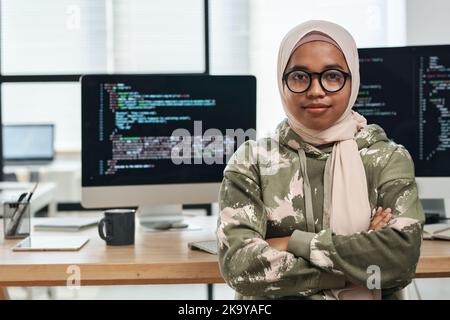 Young confident female IT engineer in hijab looking at camera while sitting by workplace against computer screens with coded data Stock Photo
