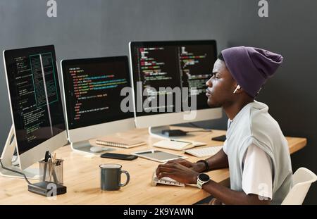 Young serious IT engineer in casualwear looking at coded data on computer screens while typing on keyboard by workplace Stock Photo