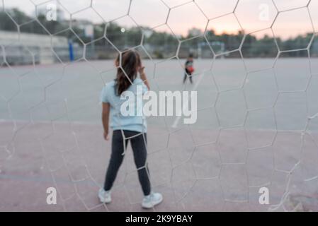 Blurred image. Back view of female child goalkeeper ready to catch a soccer ball stand on soccer field in football goal. Selective focus on girl. Stock Photo