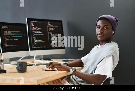 Young black man in casualwear keeping hands on computer keyboard while sitting in front of monitors and looking at camera Stock Photo