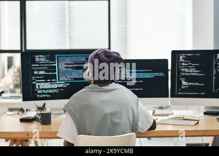 Back view of young software developer sitting by workplace in front of three computer monitors with coded data on screens Stock Photo